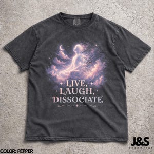 May include: Pepper-colored t-shirt featuring a celestial design with a figure in a meditative pose, surrounded by clouds and a crescent moon. The shirt has the words "LIVE, LAUGH, DISSOCIATE" in a stylized font. The brand logo "J&S Essential" is in the bottom right corner.