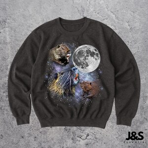 Puede incluir: Sudadera gris oscuro con un gráfico de tres castores sobre un fondo estrellado y una luna llena. Los castores están en varias poses, uno de ellos parece aullar. La sudadera tiene cuello redondo y mangas largas. La marca "J&S Essential" está impresa en la parte inferior derecha.