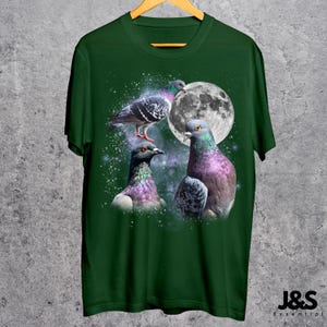 Puede incluir: Camiseta verde oscuro con un dise&ntilde;o de tres palomas con plumas iridiscentes, una luna llena y un fondo estrellado. La camiseta tiene cuello redondo y mangas cortas. La marca "J&S Essential" est&aacute; impresa en blanco en la parte inferior derecha.