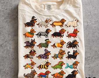 Può includere: Una t-shirt color avorio con una stampa grafica di ventiquattro bassotti vestiti con vari costumi di Halloween, tra cui una zucca, un fantasma, una mummia, un vampiro, uno scheletro, un ragno, un'ape, una sirena e un supereroe, disposti in file.