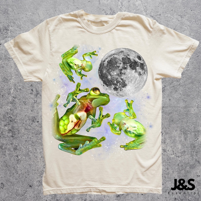 May include: Cream-colored t-shirt featuring a whimsical design of three green frogs and a grayscale moon. The frogs are in various poses, with a starry background. The shirt has a crew neck and short sleeves. The brand name "J&S Essential" is printed at the bottom right.