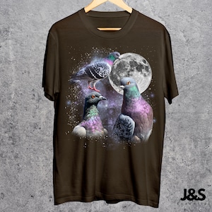 Puede incluir: Camiseta marr&oacute;n con un dise&ntilde;o de tres palomas coloridas frente a una luna llena. Las aves tienen plumas iridiscentes en tonos morados, verdes y azules. La camiseta est&aacute; colgada en una percha de madera.