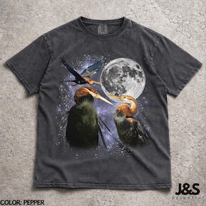 Peut inclure: T-shirt couleur poivre avec un motif de deux oiseaux au long bec, une pleine lune et un oiseau en vol. Le motif est placé sur un fond étoilé. Le texte "J&S Essential" est en bas à droite.