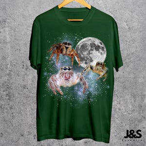 May include: Green t-shirt featuring a design of three jumping spiders with a full moon in the background. The spiders are brown and white, with detailed textures. The shirt has the text "J&S Essential" at the bottom.