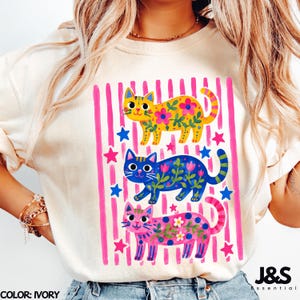 Può includere: Una t-shirt color avorio con una stampa grafica di tre gatti in giallo, blu e rosa. Ogni gatto è decorato con motivi floreali e viticci. Lo sfondo della grafica è composto da strisce verticali rosa e piccole stelle blu e rosa.