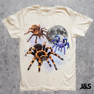May include: Cream-colored t-shirt featuring a graphic design of three spiders and a full moon. The spiders are brown, black, and blue, with detailed textures. The moon is gray and white. The shirt has a classic crew neck and short sleeves.
