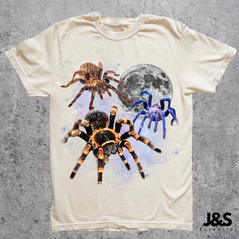 May include: Cream-colored t-shirt featuring a graphic design of three spiders and a full moon. The spiders are brown, black, and blue, with detailed textures. The moon is gray and white. The shirt has a classic crew neck and short sleeves.