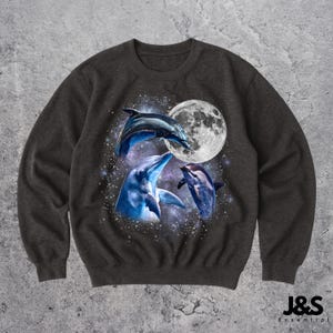 Peut inclure: Sweat-shirt gris foncé avec un motif de trois dauphins sautant dans un espace étoilé avec une pleine lune. Les dauphins sont dans des tons de bleu et de blanc. Le sweat-shirt a un col rond et des manches longues. La marque "J&S Essential" est imprimée en blanc.