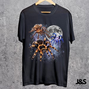 May include: Black t-shirt featuring a graphic of three spiders in front of a full moon and starry background. The spiders are brown, orange, and blue. The shirt has the text "J&S Essential" at the bottom.
