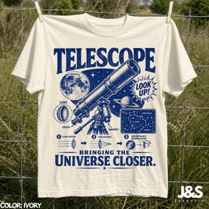 May include: An ivory t-shirt featuring a blue graphic print of a telescope, the moon, Saturn, and a star chart. The text reads "TELESCOPE", "LOOK UP!", and "BRINGING THE UNIVERSE CLOSER." It includes diagrams explaining lens focus, light entry, and magnification.