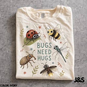 Può includere: Una t-shirt color avorio con una stampa grafica raffigurante una coccinella su una foglia, un bombo, una libellula, uno scarabeo e una lucciola. Il testo BUGS NEED HUGS è stampato al centro in lettere verde acqua, circondato da piccole stelle.