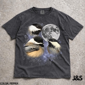 Pode incluir: Camiseta cinza escuro com uma estampa de três chapins, uma lua cheia e um céu estrelado. A camiseta tem um aspecto desbotado. O texto "J&S Essential" está impresso no canto inferior direito.