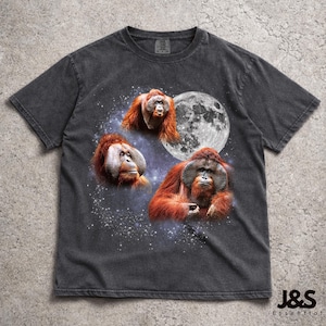 Puede incluir: Camiseta gris oscuro con un diseño de tres orangutanes sobre un fondo estrellado y una luna llena. Los orangutanes tienen pelaje marrón rojizo y están en diferentes poses. La camiseta tiene un aspecto vintage.