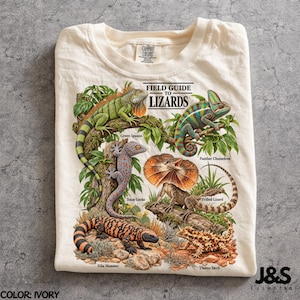 Puede incluir: Una camiseta color marfil con un estampado gráfico titulado FIELD GUIDE TO LIZARDS. El diseño muestra una iguana verde, un camaleón pantera, un gecko tokay, un lagarto de collar, un monstruo de Gila y un diablo espinoso, etiquetados en negro.