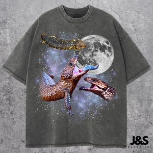 May include: Dark gray t-shirt featuring a graphic of three lizards in space with a full moon. The lizards are brown and gold with blue tongues. The text "J&S Essential" is at the bottom right.
