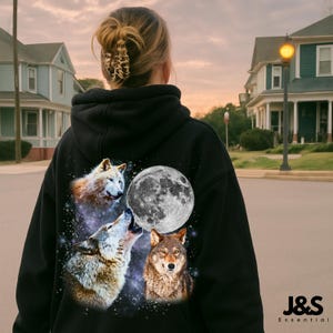 May include: Black hooded sweatshirt with a graphic of three wolves and a full moon against a starry backdrop. The wolves are in shades of brown, white, and grey. The sweatshirt has a drawstring hood and a kangaroo pocket.