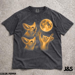 Puede incluir: Camiseta gris oscuro con un diseño de tres zorros y un gato mirando una luna llena. El diseño es en tonos naranja y amarillo. El texto "J&S Essential" está en la parte inferior, y la camiseta es color pimienta.