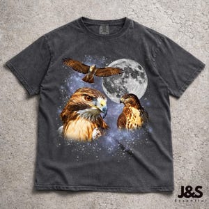 May include: Dark grey t-shirt with a detailed graphic of hawks, a full moon, and a starry night sky. The design includes three hawks in various poses, with the moon in the background. The shirt has a vintage, worn appearance.