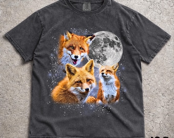 May include: Dark gray t-shirt featuring a graphic of three foxes and a full moon. The foxes are depicted in shades of orange and white, with a starry background. The text "J&S Essential" is at the bottom right.
