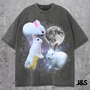 Peut inclure: Un t-shirt gris foncé avec un imprimé graphique de trois lapins blancs et une lune. Les lapins sont dans des poses différentes, et la lune est en arrière-plan.