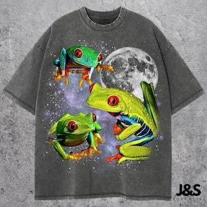 May include: Dark gray t-shirt featuring a vibrant graphic of three red-eyed tree frogs against a starry night sky and a full moon. The frogs are in shades of green, yellow, and blue. The text "J&S Essential" is at the bottom.