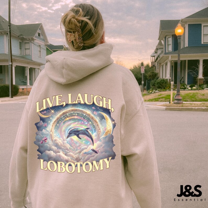 May include: Beige hoodie with the words "LIVE, LAUGH, LOBOTOMY" printed on the back. The graphic features a dolphin jumping through a colorful, celestial design with clouds and crescent moons. The hoodie has a drawstring hood.
