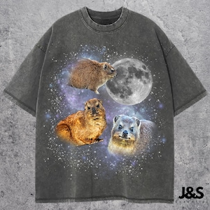 Puede incluir: Una camiseta gris con un estampado descolorido de tipo galaxia con tres animales hyrax de roca. Los hyrax son marrones y grises y están posicionados sobre un fondo de estrellas y una luna creciente.