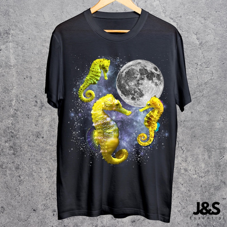 Może przedstawiać: Czarny t-shirt z trzema ż&oacute;łtymi konikami morskimi, pełnią księżyca i gwiezdnym tłem. Koniki morskie mają ż&oacute;łte i zielone kolory. Koszulka wisi na drewnianym wieszaku.