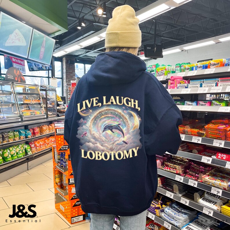 May include: Navy blue hoodie with the text "LIVE, LAUGH, LOBOTOMY" and a graphic of a dolphin jumping through a colorful, celestial design. The hoodie is oversized and has a hood and long sleeves. A yellow beanie is worn.
