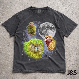 Puede incluir: Camiseta gris oscuro con un gráfico de tres pájaros coloridos, una luna llena y un fondo estrellado. Los pájaros son en tonos verdes, amarillos y rojos. La camiseta tiene un aspecto vintage y desgastado. El logo de la marca "J&S Essential" está en la parte inferior derecha.