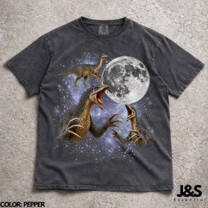 Op de afbeelding: Pepperkleurig t-shirt met een gedetailleerde illustratie van dinosaurussen met open monden, een volle maan en een sterrenachtergrond. Het shirt bevat onderaan de tekst "J&S Essential".