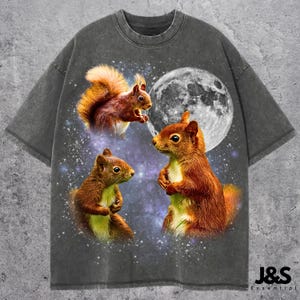 May include: Gray t-shirt featuring a whimsical design of three squirrels against a starry night sky with a full moon. The squirrels are depicted in shades of brown and orange, with the text "J&S Essential" at the bottom.