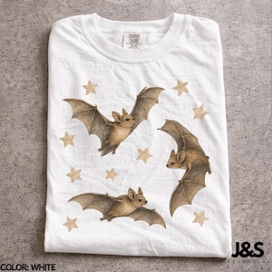 May include: White t-shirt featuring a watercolor design of three bats with outstretched wings and scattered star shapes. The bats are brown with detailed wings. The shirt has a crew neck and short sleeves.