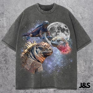 May include: Dark grey t-shirt with a graphic of three iguanas, a full moon, and a starry background. The shirt has a vintage wash appearance. The text "J&S Essential" is printed in the lower right corner.