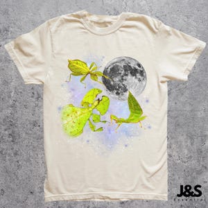 May include: Cream-colored t-shirt featuring a whimsical design. The graphic includes a full moon, leaf insects, and a starry background. The shirt has a classic crew neck and short sleeves. The brand logo is in the bottom right corner.