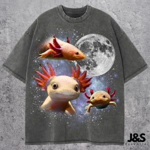May include: Dark grey t-shirt with a graphic of three axolotls, a full moon, and a starry background. The axolotls are orange, yellow, and green. The text "J&S Essential" is at the bottom right.
