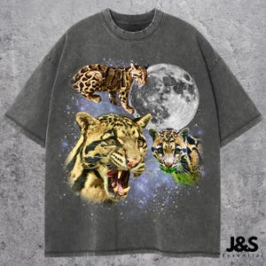 Op de afbeelding: Donkergrijs t-shirt met een grafisch ontwerp van drie nevelpanters, een volle maan en een sterrenhemel. Het shirt heeft een ronde hals en korte mouwen. De merknaam "J&S Essential" staat rechtsonder in het wit gedrukt.