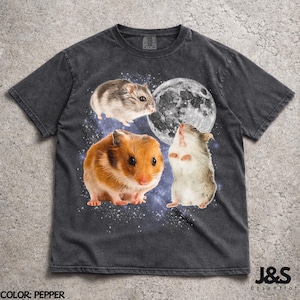 Könnte beinhalten: Ein anthrazitfarbenes T-Shirt mit einem Grafikdruck von drei Hamstern in einer sternenklaren Weltraumumgebung mit einem großen Mond. Ein Hamster ist braun, einer ist grau-weiß und einer steht auf seinen Hinterbeinen. Das T-Shirt hat einen Rundhalsausschnitt.
