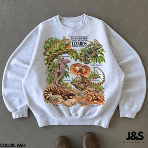 May include: An ash grey crewneck sweatshirt featuring a graphic print titled Field Guide to Lizards. The illustration displays a green iguana, panther chameleon, tokay gecko, frilled lizard, Gila monster, and thorny devil with labels.