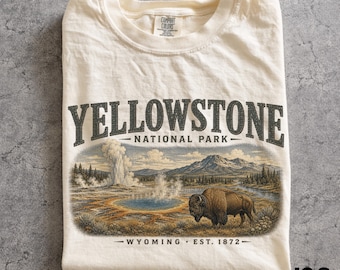 May include: Ivory-colored t-shirt featuring a graphic of Yellowstone National Park. The design includes a geyser, lake, mountains, and a bison. Text on the shirt reads "YELLOWSTONE NATIONAL PARK - WYOMING EST. 1872-".