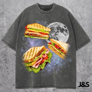 May include: Dark gray t-shirt featuring a graphic of three sandwiches floating in space with a full moon. The sandwiches have various fillings, including ham, cheese, lettuce, and tomato. The shirt has the text "J&S Essential" at the bottom.