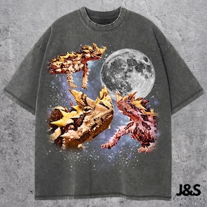 Pode incluir: Camiseta cinza escuro com um design gráfico de três demônios espinhosos, uma lua cheia e um fundo estrelado. A camiseta tem um visual vintage e desbotado. O logotipo da marca "J&S Essential" está na parte inferior direita.