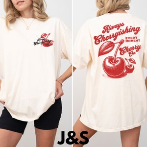 May include: A white oversized t-shirt with a red cherry graphic and the text "Always Cherryishing Every Moment Cherry On" on the back. The front of the shirt has a smaller version of the cherry graphic and the text "Always Cherryishing Every Moment".
