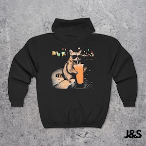 May include: A black hooded sweatshirt featuring a graphic print on the back of a raccoon wearing black sunglasses and holding an orange drink with a straw. The raccoon is positioned against a dark background with blurred colorful city lights.