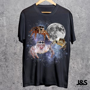 May include: Black t-shirt featuring a design of three jumping spiders with a full moon and starry background. The spiders are in shades of brown, tan, and white. The shirt is hanging on a wooden hanger.