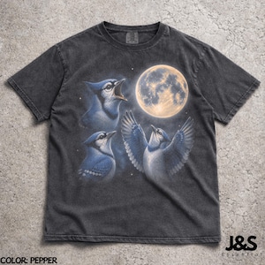 May include: Pepper-colored t-shirt featuring a detailed illustration of three blue jays with a full moon in the background. The birds are depicted in various poses, with one singing, another looking up, and one with outstretched wings. The text "J&S Essential" is in the lower right corner.