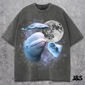 May include: Dark gray t-shirt featuring a graphic of three beluga whales, a full moon, and a starry background. The shirt has a relaxed fit and a crew neck. The text "J&S Essential" is printed on the bottom right.