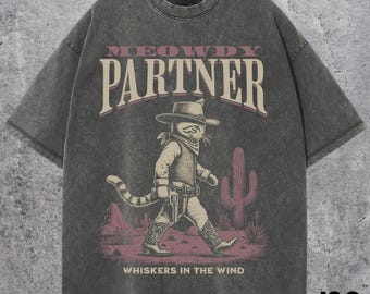 Pode incluir: T-shirt cinzenta com um desenho de gato cowboy, com o texto "MEOWDY PARTNER" e "WHISKERS IN THE WIND". O design tem um aspeto vintage e desgastado. A t-shirt tem um corte folgado.