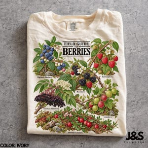 May include: An ivory t-shirt featuring a botanical illustration titled FIELD GUIDE TO BERRIES. The graphic displays clusters of highbush blueberries, red raspberries, blackberries, American elderberries, gooseberries, and American cranberries.