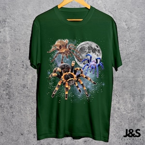May include: Green t-shirt featuring a graphic of three spiders in front of a full moon and a starry background. The spiders are brown, black, and blue. The shirt has a crew neck and short sleeves. The brand name "J&S Essential" is at the bottom right.